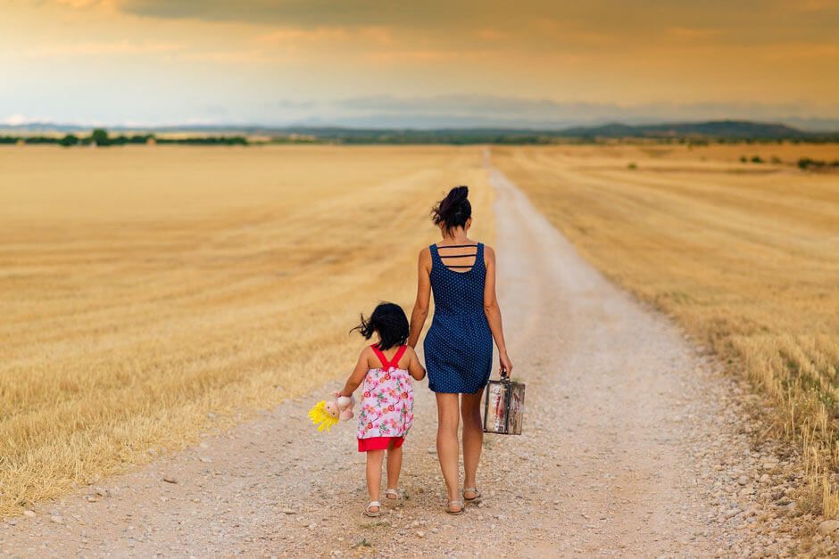 A mother and her child, who is holding a doll, walk down a dirt road